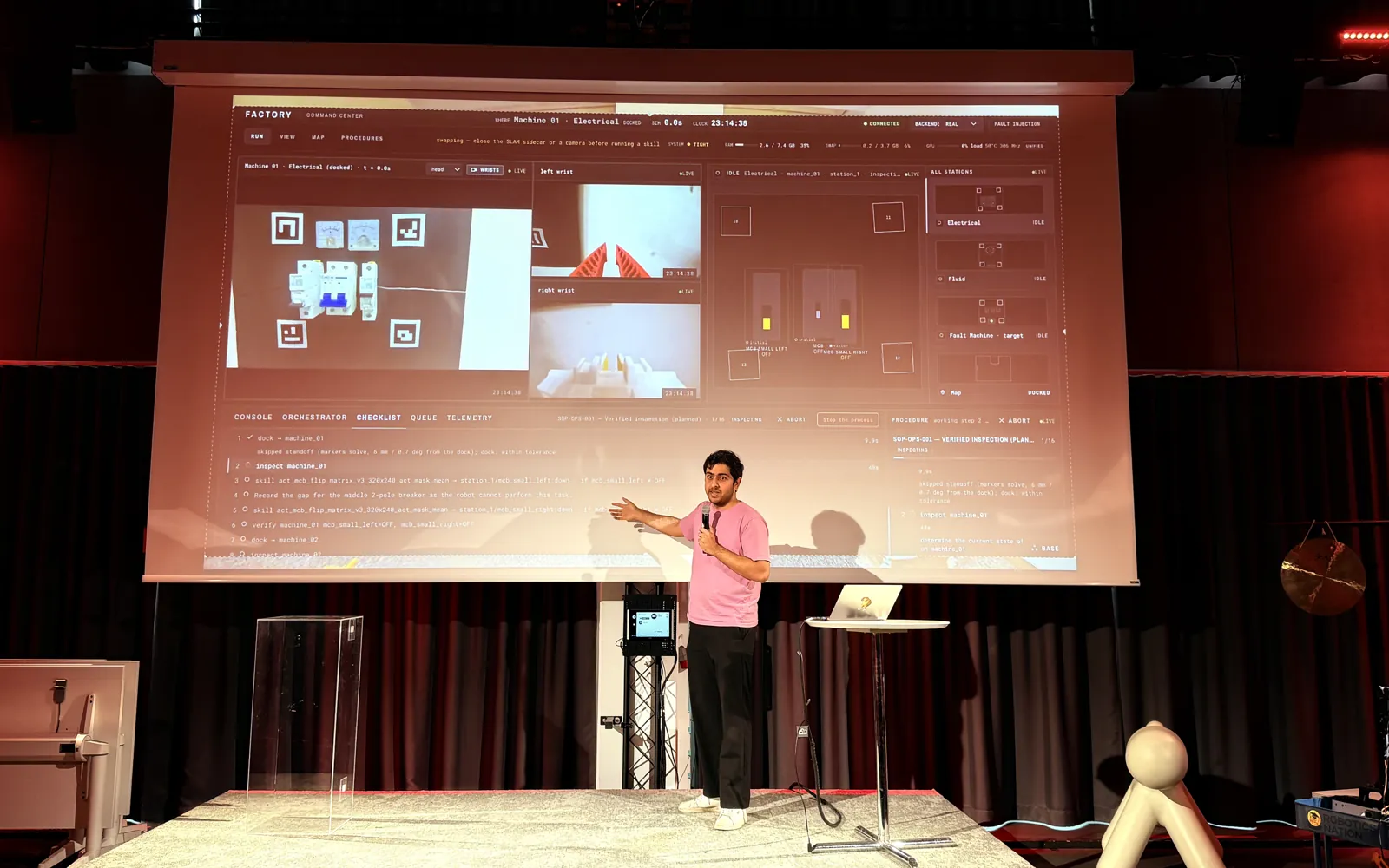 Zuper presenting on stage in front of his Factory command center interface, showing the breaker panel camera with fiducial markers, both wrist cameras and the skill checklist